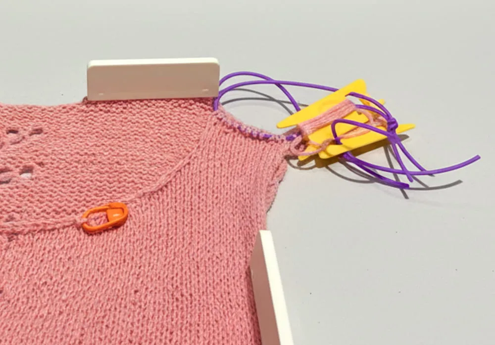 Pink hand-knitted fabric for a knitting project, featuring a stitch marker, yarn bobbin with yarn, and knitting needle holders