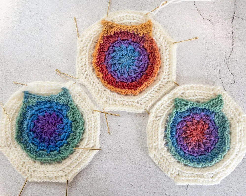 Three colorful crocheted owl motifs laid flat with bobby pins used as stitch markers, each with cream outer rounds and bright circular centers.