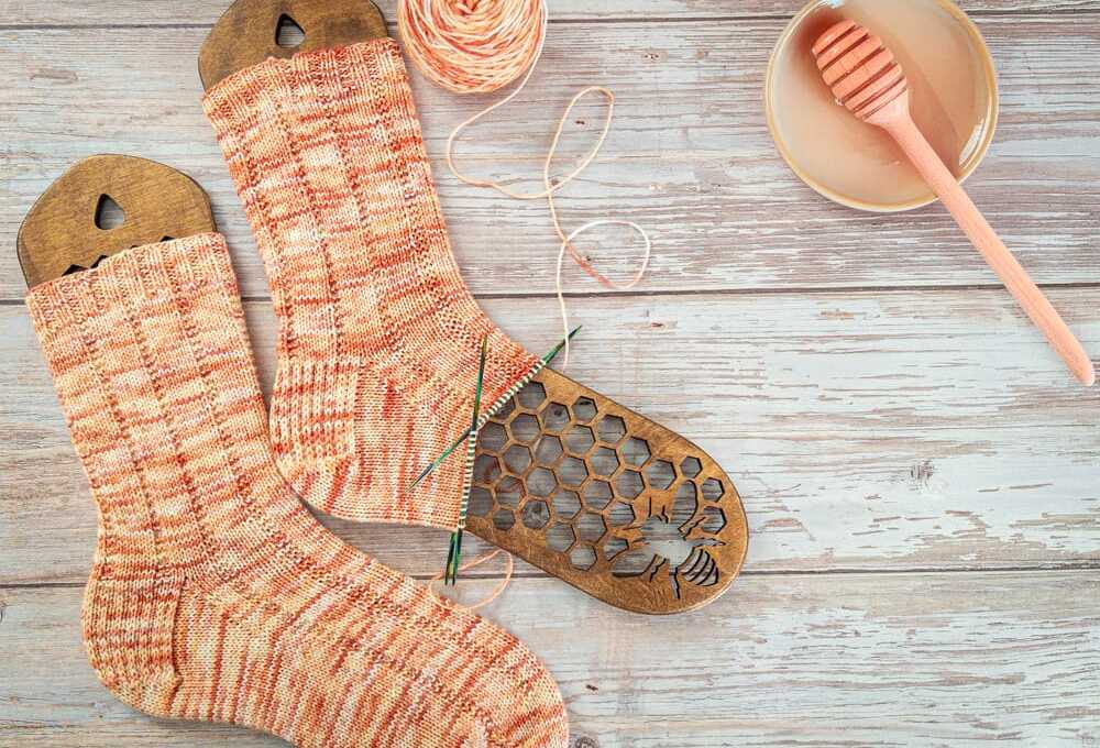 Peach and cream hand-knit socks, one still in progress, shown with matching yarn on wooden sock blockers with a honeycomb and bee design