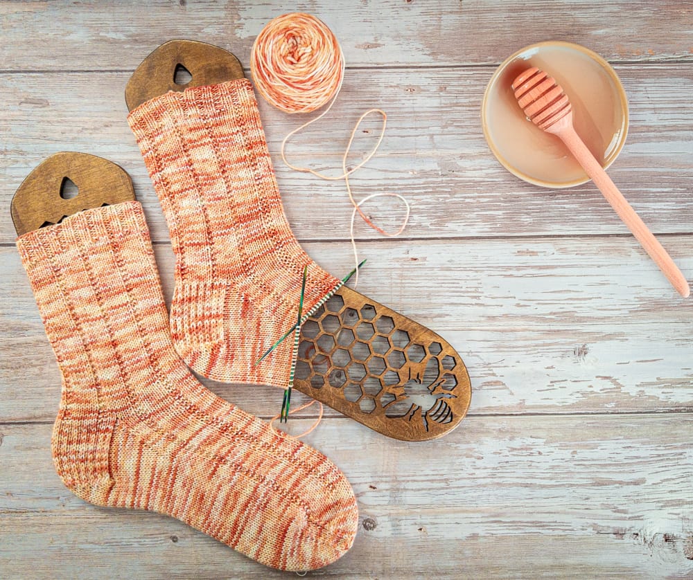 Peach and cream hand-knit socks, one still in progress, shown with matching yarn on wooden sock blockers with a honeycomb and bee design