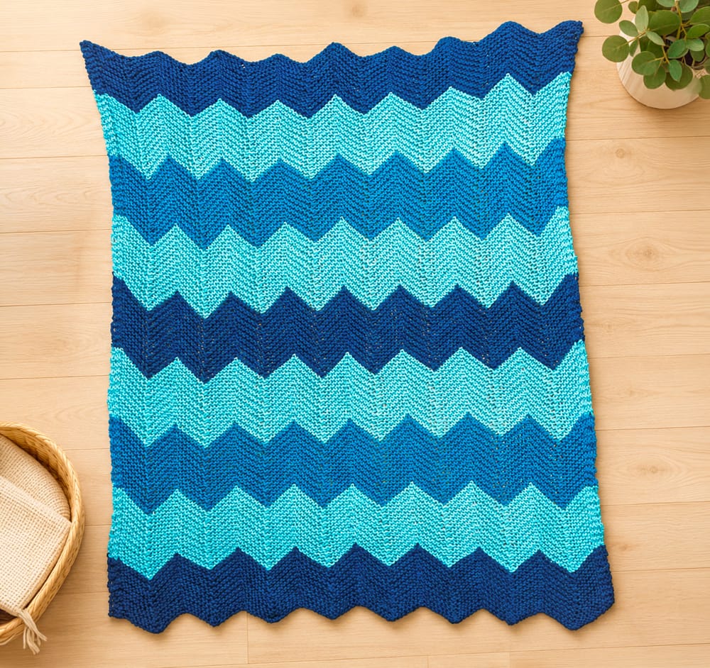 Chevron-knit baby blanket laid flat on a light wood floor with alternating turquoise and navy zigzag stripes, in garter-stitch. There is a small potted plant at the top-right and a woven basket at the bottom-left, just to be cute.