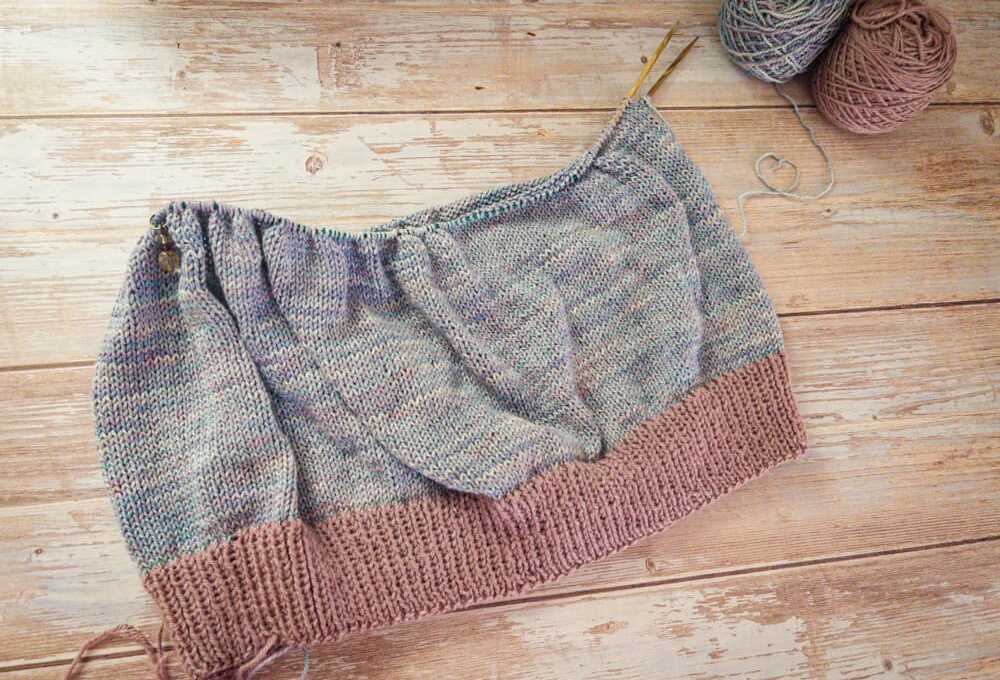 A knitted top in progress laid flat on a pale wood background, with a mauve ribbed hem and a speckled gray-blue body on circular needles. Two balls of yarn sit in the upper corner, one in the main speckled color and one in the contrasting mauve.