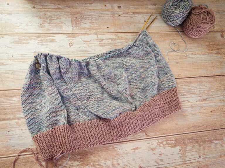 A knitted top in progress laid flat on a pale wood background, with a mauve ribbed hem and a speckled gray-blue body on circular needles. Two balls of yarn sit in the upper corner, one in the main speckled color and one in the contrasting mauve.