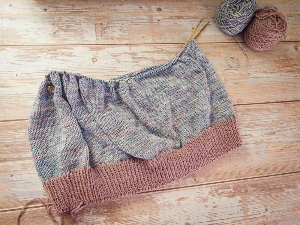 A knitted top in progress laid flat on a pale wood background, with a mauve ribbed hem and a speckled gray-blue body on circular needles. Two balls of yarn sit in the upper corner, one in the main speckled color and one in the contrasting mauve.