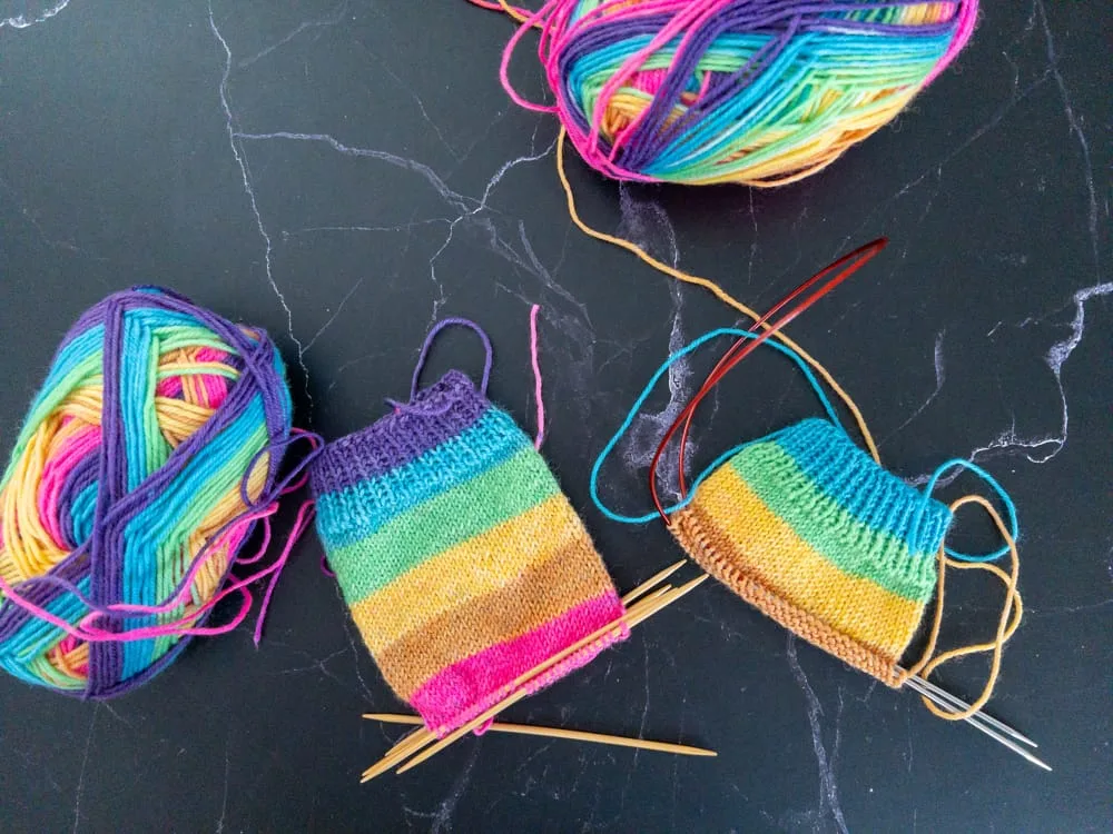 Two small striped socks in progress on a dark surface, knit from Knit Picks Felici yarn in the Lisa colorway. The socks are in rainbow stripes in purple, teal, green, yellow, tan, and pink, with balls of yarn and needles spread around them. One sock starts with a purple strip and one with blue.