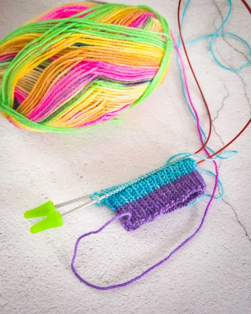 The beginning of a sock cuff in progress on circular needles, knit from Knit Picks Felici yarn in the Lisa colorway. A ball of the brightly striped yarn sits nearby, showing neon green, yellow, orange, pink, teal, and purple sections.