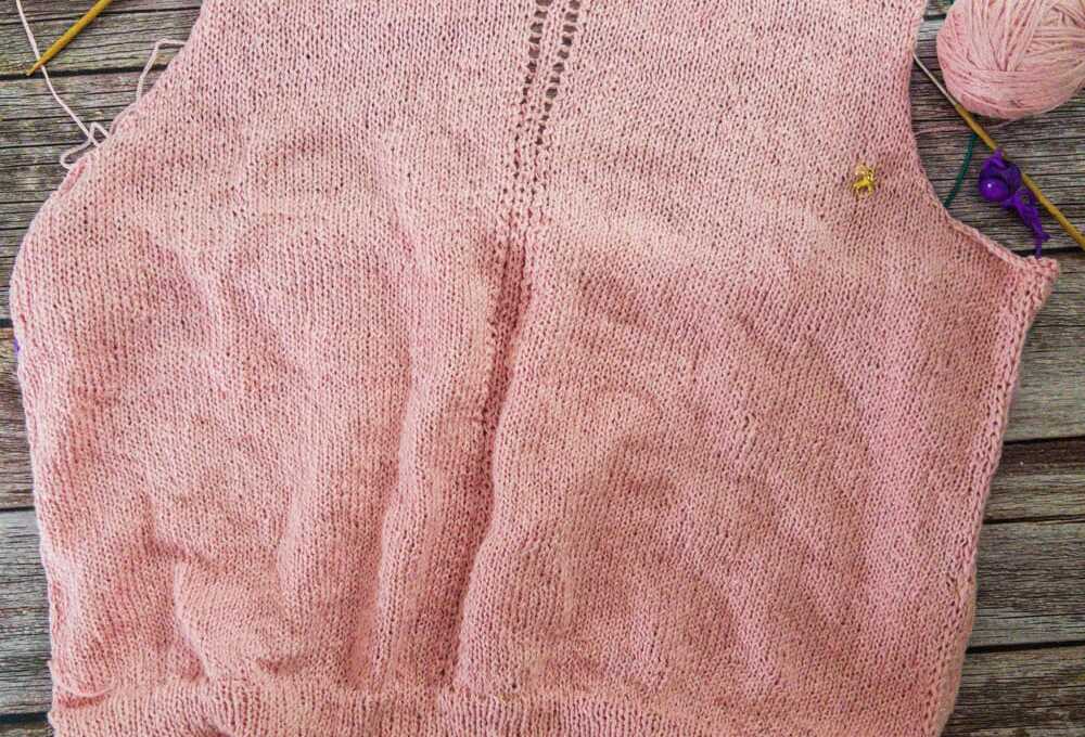 The back of a pale pink knitted top in progress laid flat on a rustic gray wood background. The piece has ribbing at the hem, smooth stockinette fabric, and a decorative lace panel running up the center back, with needles, yarn, and stitch markers visible around the edges.