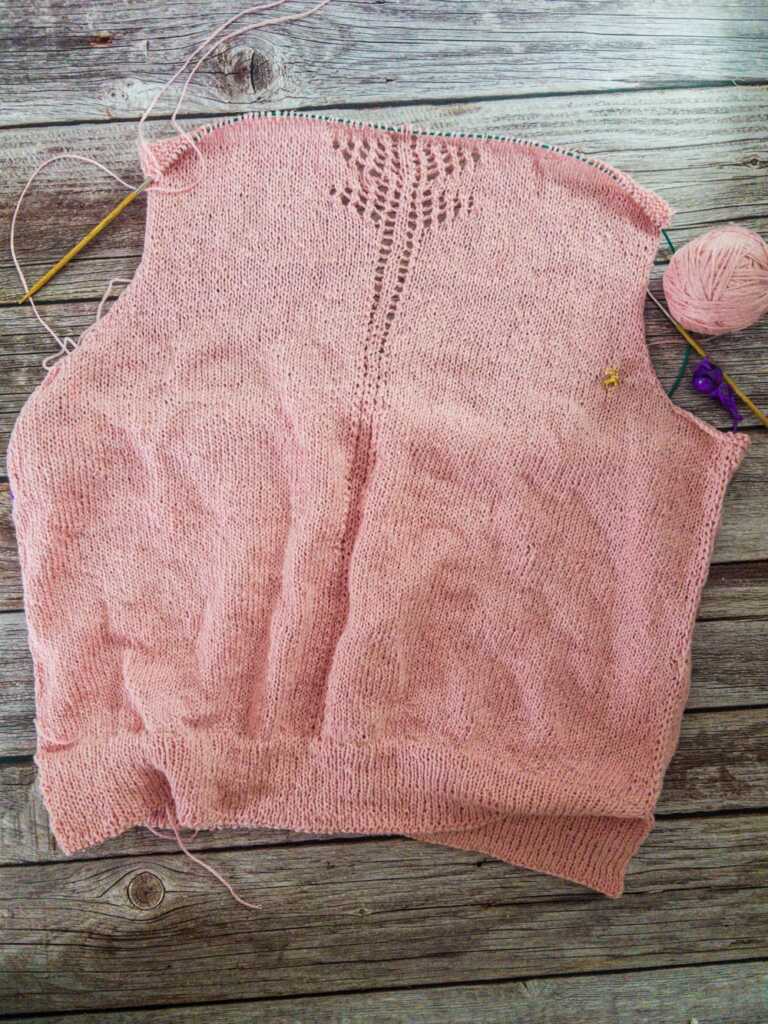 The back of a pale pink knitted top in progress laid flat on a rustic gray wood background. The piece has ribbing at the hem, smooth stockinette fabric, and a decorative lace panel running up the center back, with needles, yarn, and stitch markers visible around the edges.