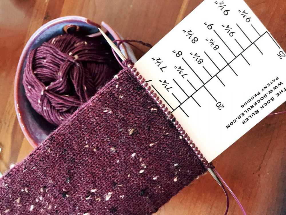 Hand-knit purple sock in progress measured with a Sock Ruler, with matching tweed yarn in the background.