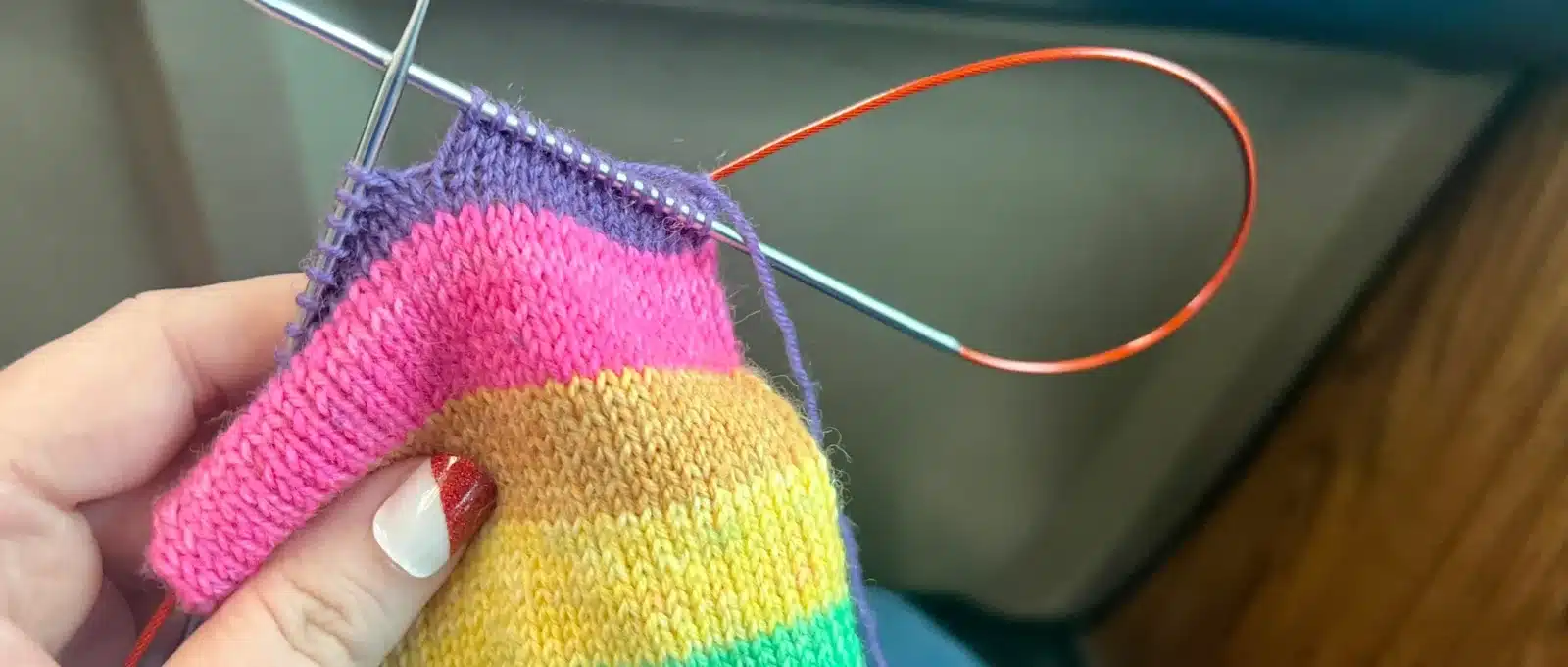 A close-up of a sock in progress knit from Knit Picks Felici yarn in the Lisa colorway, with bright stripes of teal, green, yellow, tan, pink, and purple. The sock is on circular needles and being held in one hand with a cool diagonal stripe of red glitter on the thumbnail.