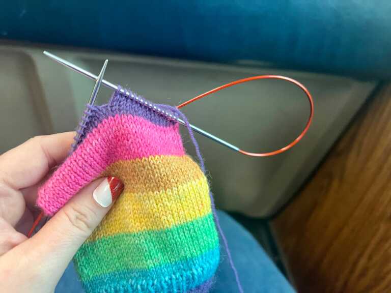 A close-up of a sock in progress knit from Knit Picks Felici yarn in the Lisa colorway, with bright stripes of teal, green, yellow, tan, pink, and purple. The sock is on circular needles and being held in one hand with a cool diagonal stripe of red glitter on the thumbnail.