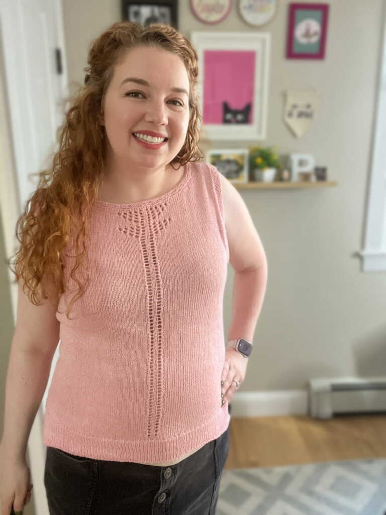 Smiling woman models the Take Wing Shell - a beautiful pink hand-knit sleeveless top, featuring intricate lace yarn patterns and a central lace panel