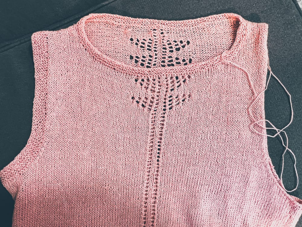 A pink hand-knitted top featuring intricate lace knitting patterns at the neckline and a central panel, a yarn craft work in progress