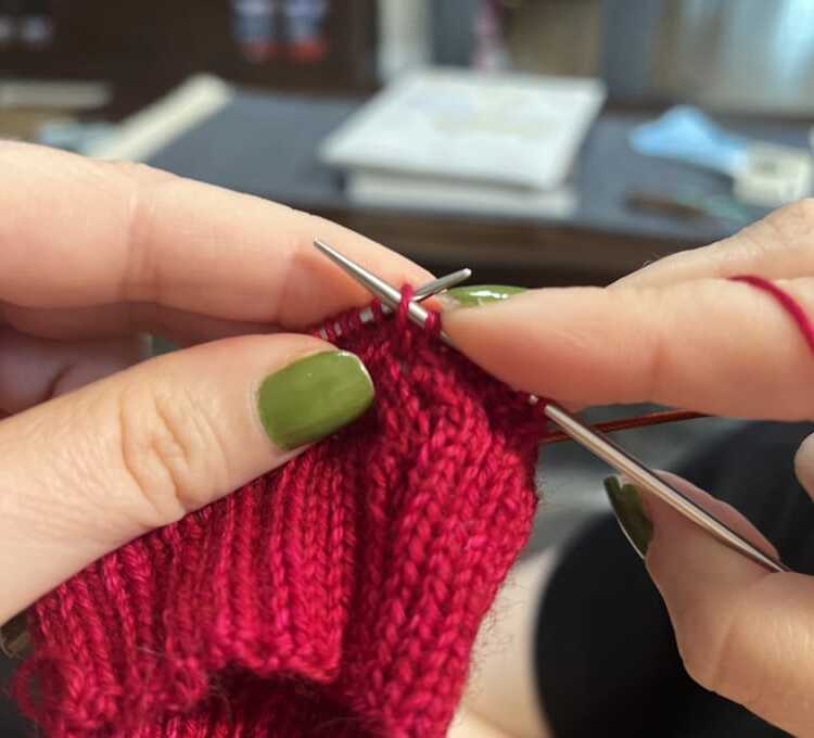 Hands with green nails knitting a handmade project using bright red yarn and metal circular needles, showing ribbed stitches