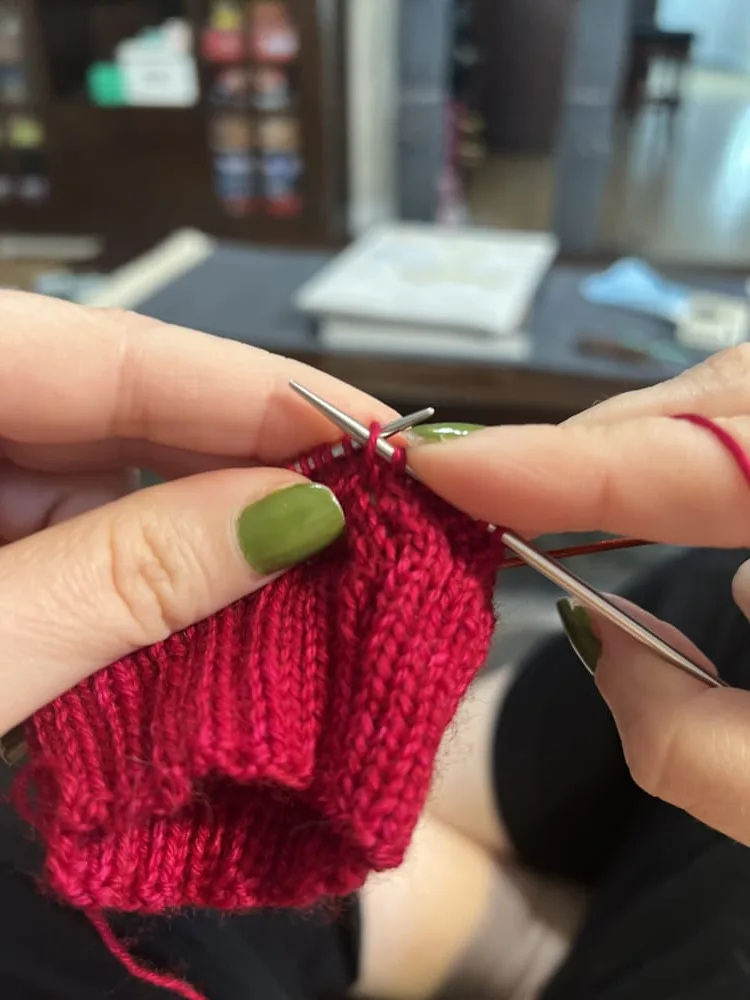 Hands with green nails knitting a handmade project using bright red yarn and metal circular needles, showing ribbed stitches