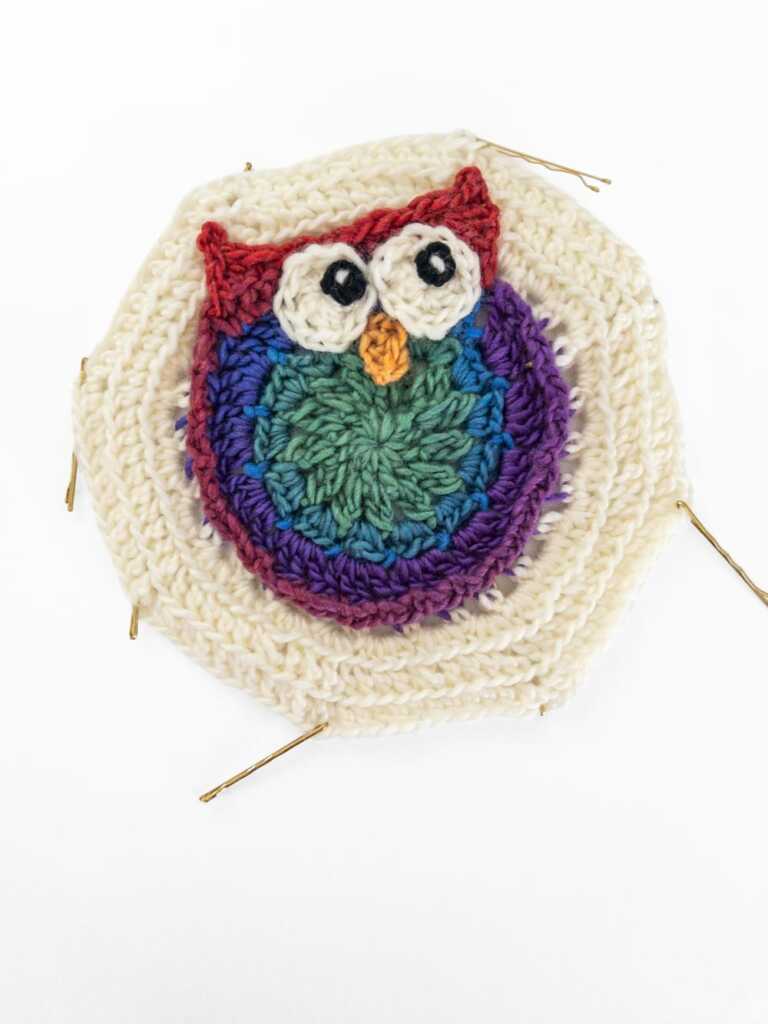 A crocheted owl granny square in rainbow yarn colors on in an octagon shape.