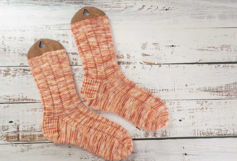 A pair of hand-knitted socks in variegated orange yarn, displayed on wooden sock blockers, showcasing a ribbed knitting pattern