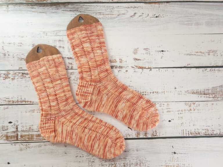 A pair of hand-knitted socks in variegated orange yarn, displayed on wooden sock blockers, showcasing a ribbed knitting pattern