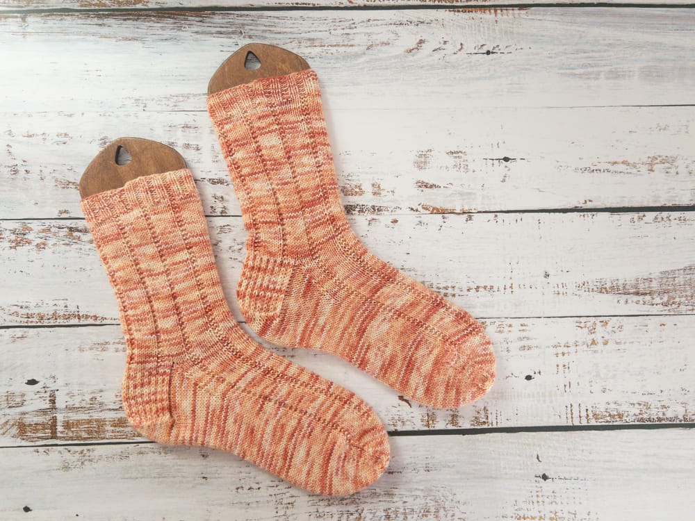 A pair of hand-knitted socks in variegated orange yarn, displayed on wooden sock blockers, showcasing a ribbed knitting pattern