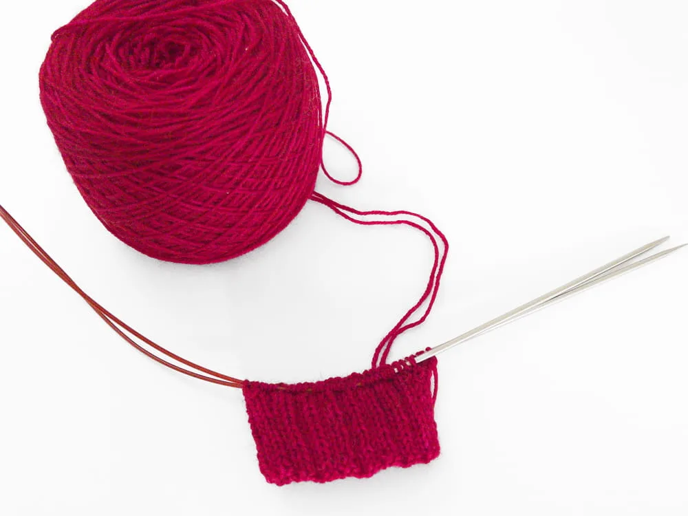 Vibrant red yarn, knitted sock cuff, and circular knitting needles.
