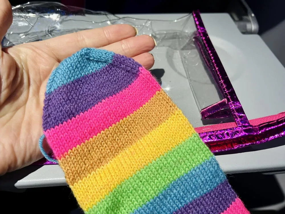 A hand holds a vibrant, rainbow self-striping yarn knitting project, showcasing colorful knit stitches