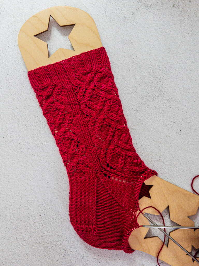 Red hand-knitted wool sock with lace and cable patterns on a wooden sock blocker with star cutouts. Knitting needles and yarn are visible