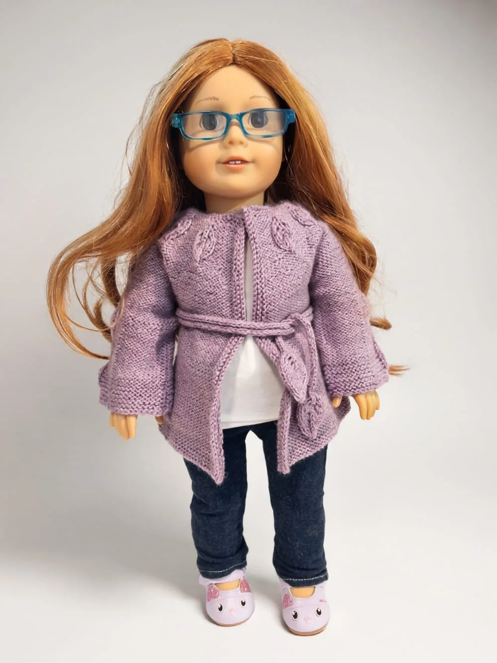 A doll wears a hand-knitted lavender cardigan with a leaf pattern, blue glasses, and jeans. Doll clothes knitting project