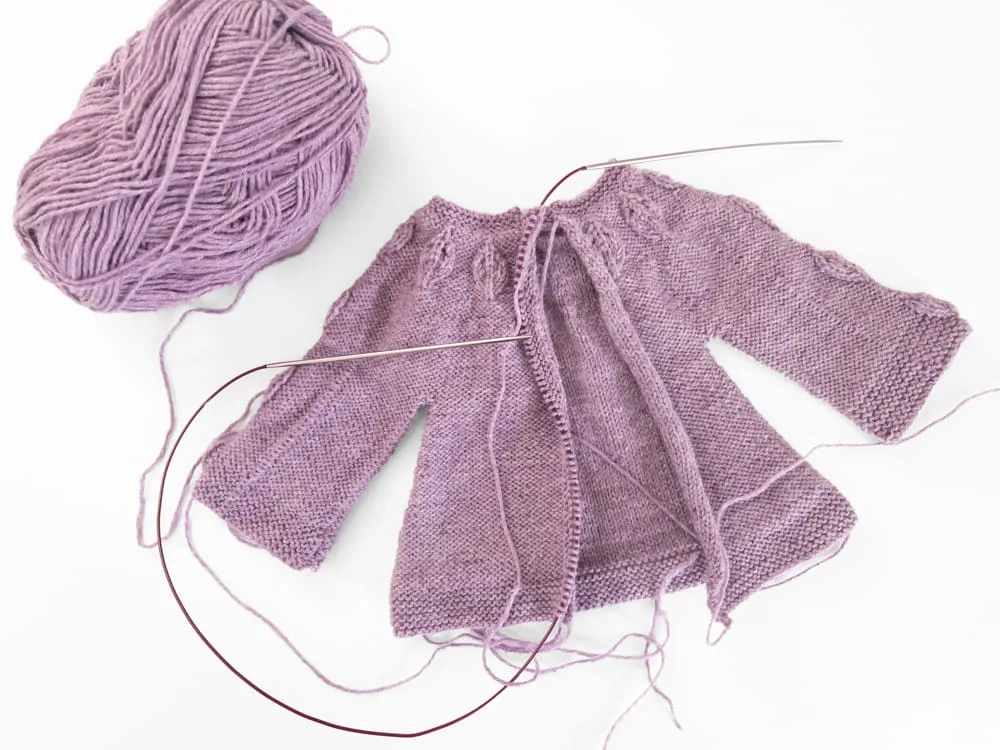 Hand-knitted purple doll cardigan sweater with the front band stiches on circular knitting needles with a ball of lavender yarn
