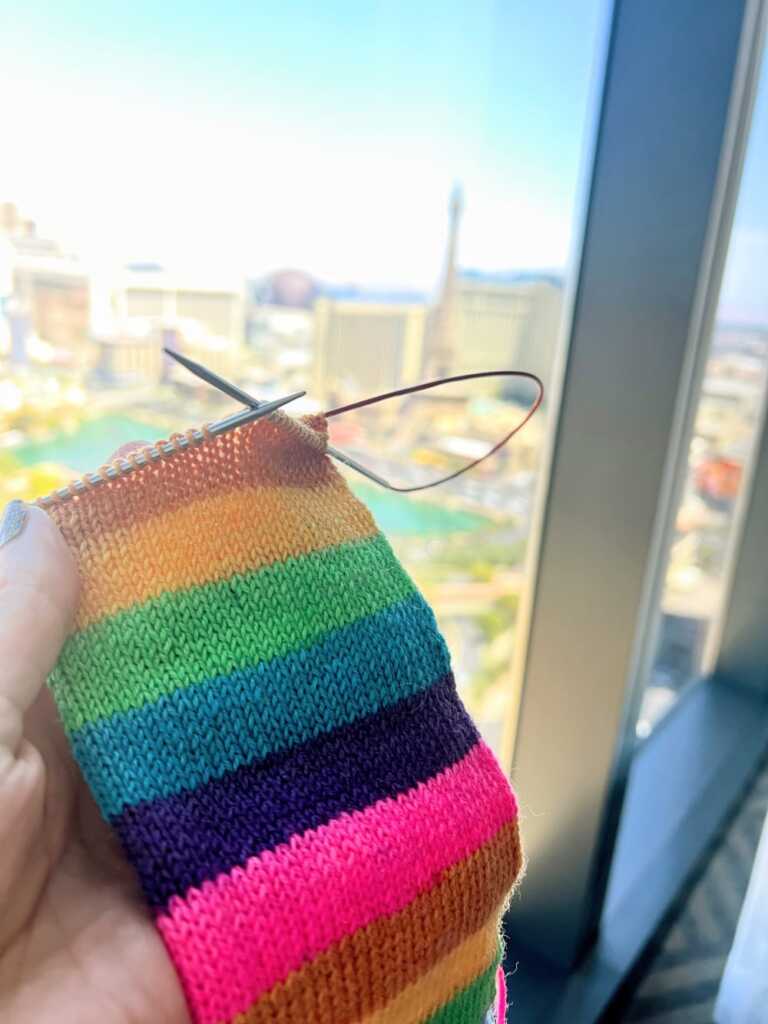 Hand knitting a vibrant rainbow striped sock project with colorful yarn on circular needles, held in front of a window overlooking the las vegas strip.
