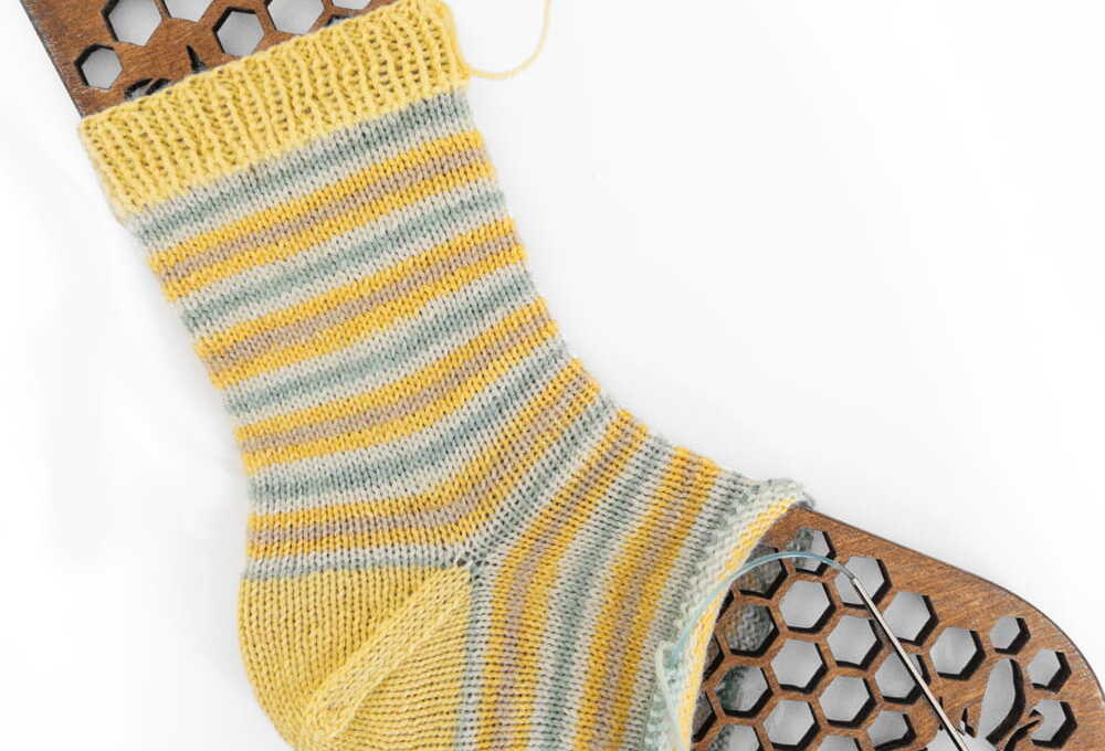 Hand-knitted striped sock in yellow and grey yarn on a wooden honeycomb sock blocker with circular knitting needles