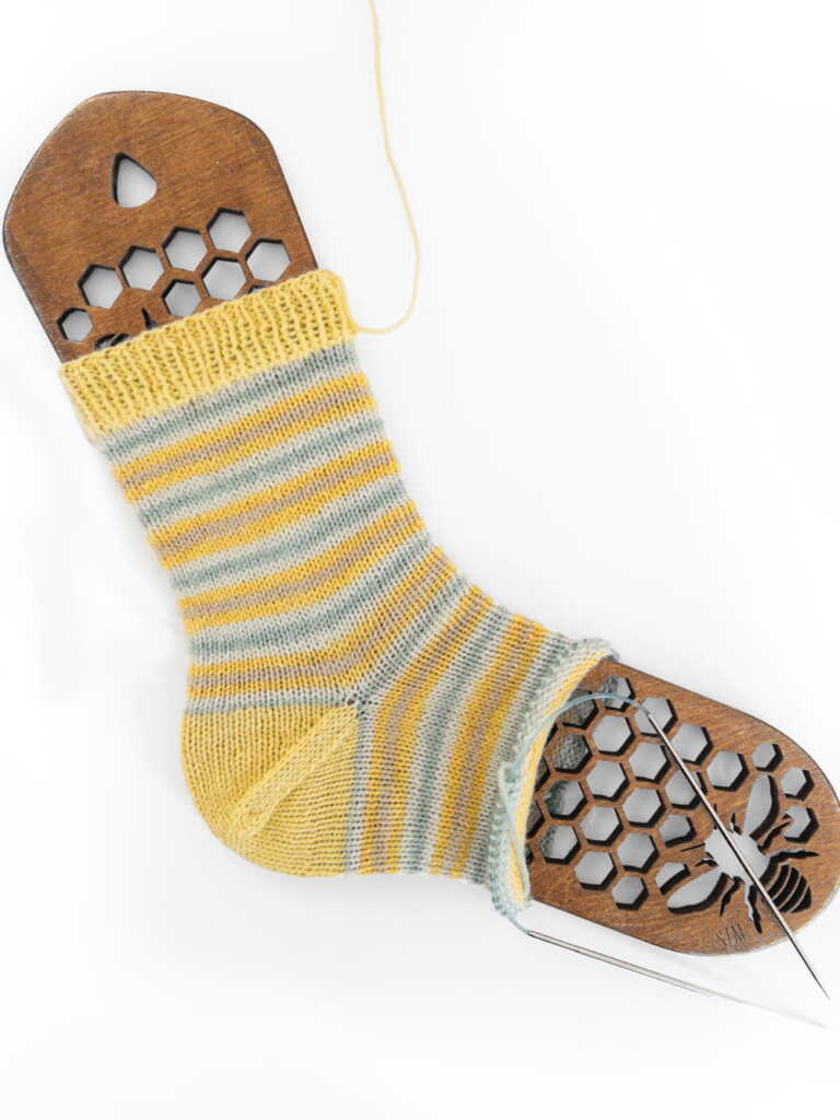 Hand-knitted striped sock in yellow and grey yarn on a wooden honeycomb sock blocker with circular knitting needles