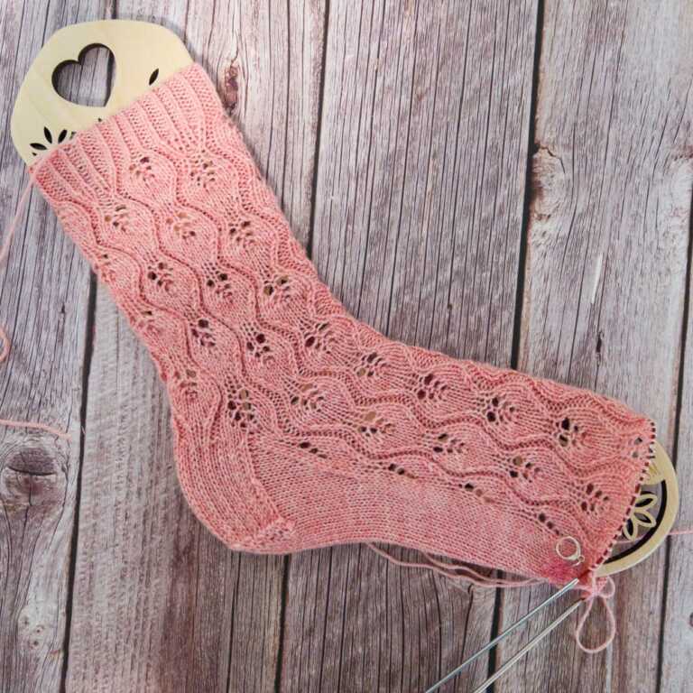Pink knitted sock with lace pattern on a sock blocker, a handmade sock knitting project with delicate yarn