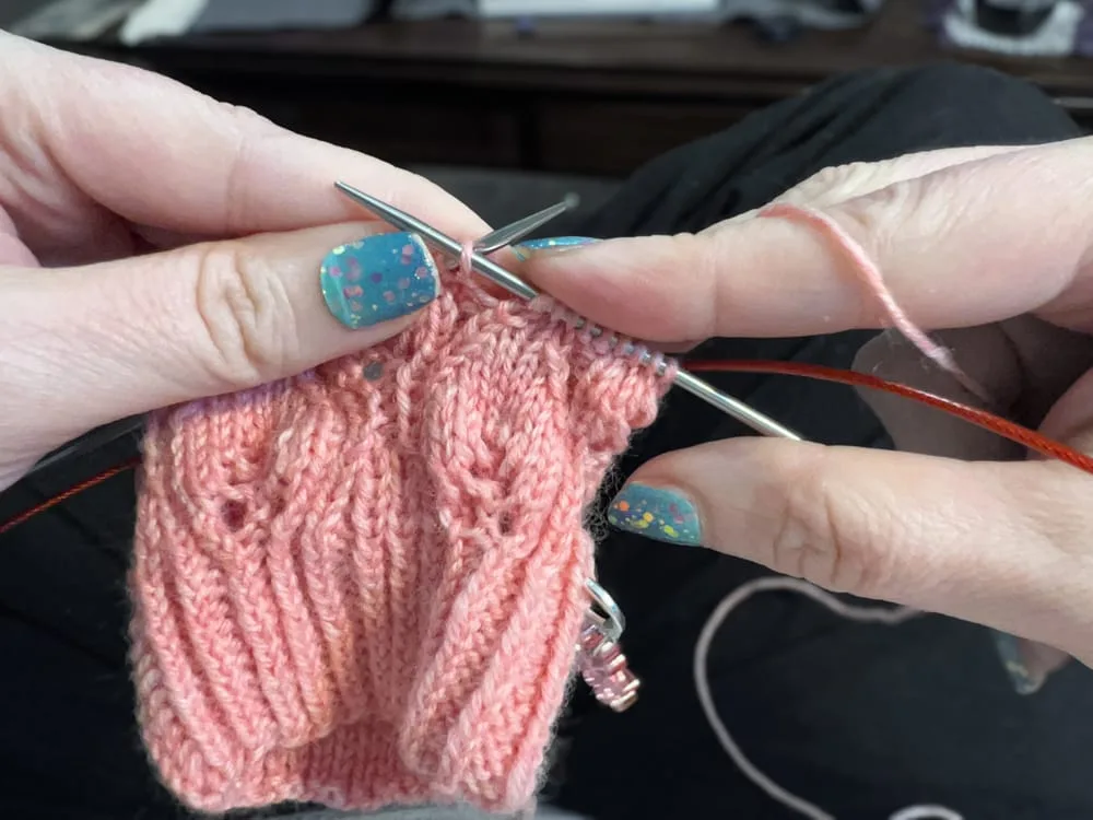 Hands using knitting needles to create a pink yarn project with a textured stitch pattern