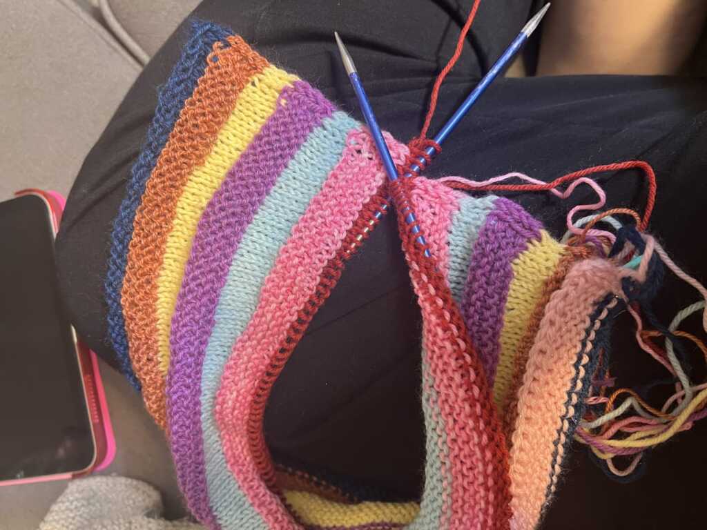 A vibrant, striped knitting project with colorful sock yarn on knitting needles. Pattern is the Mix Tape Tee by Wool and Pine.