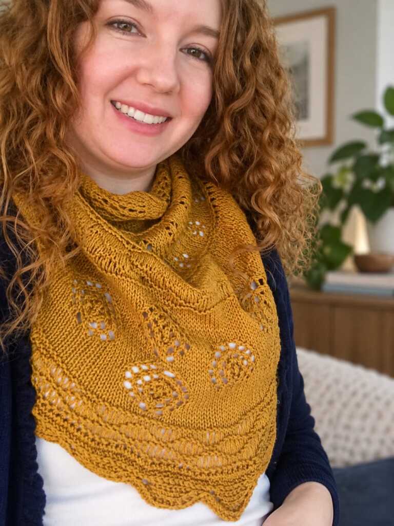 Smiling woman in a hand-knitted mustard yellow lace shawl featuring an intricate eyelet knitting pattern