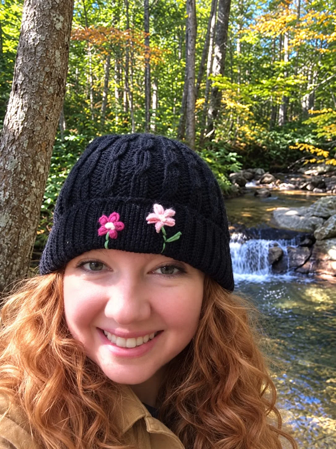 Smiling woman wearing a black cable knit beanie with embroidered flowers, a cozy knitwear design for fall yarn projects